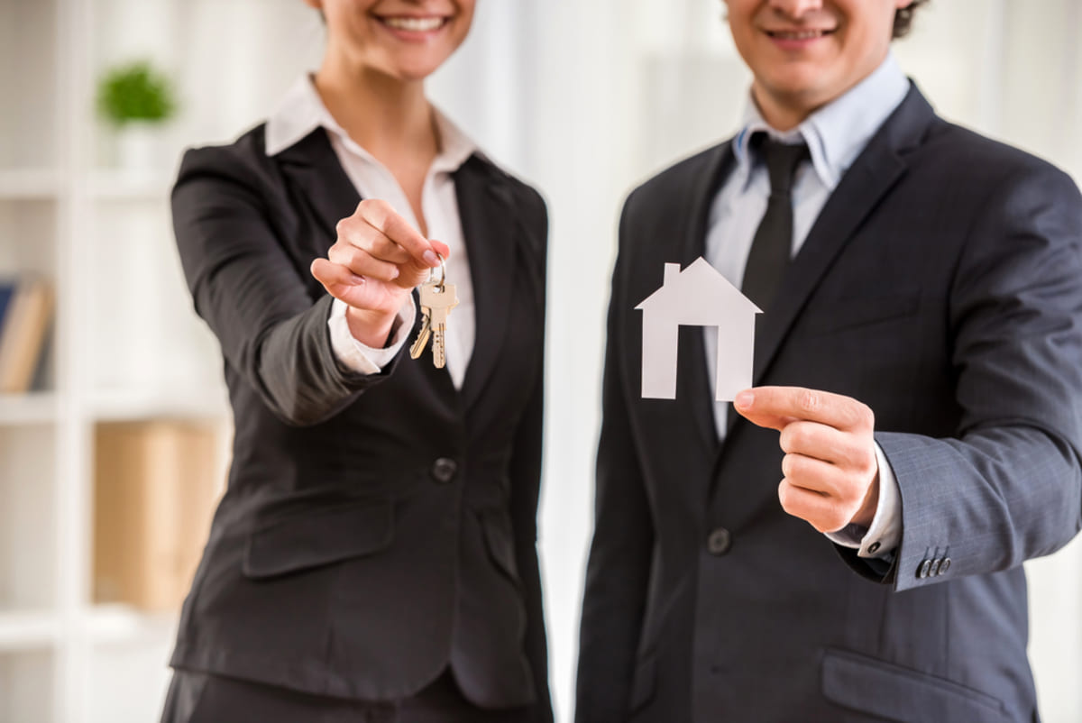A woman in a suit holding keys and a man in a suit holding a wooden house